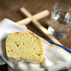 Eine Scheibe Brot liegt auf einer Stoffserviette auf einem Tisch, daneben liegt ein schlichtes Holzkreuz und steht ein Glas Wasser 