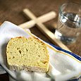 Eine Scheibe Brot liegt auf einer Stoffserviette auf einem Tisch, daneben liegt ein schlichtes Holzkreuz und steht ein Glas Wasser 