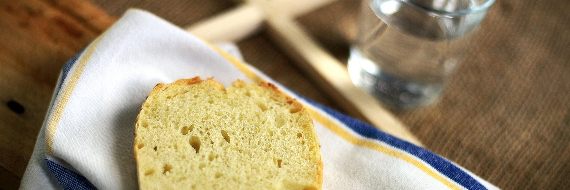 Eine Scheibe Brot liegt auf einer Stoffserviette auf einem Tisch, daneben liegt ein schlichtes Holzkreuz und steht ein Glas Wasser 