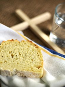 Eine Scheibe Brot liegt auf einer Stoffserviette auf einem Tisch, daneben liegt ein schlichtes Holzkreuz und steht ein Glas Wasser 