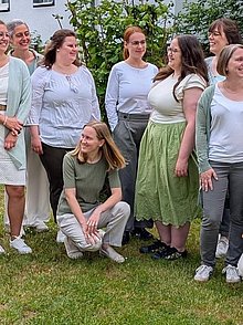 Eine Gruppe junger Frauen, die sich teilweise gegenseitig froh anblickend auf einer Wiese in einem Garten stehen.