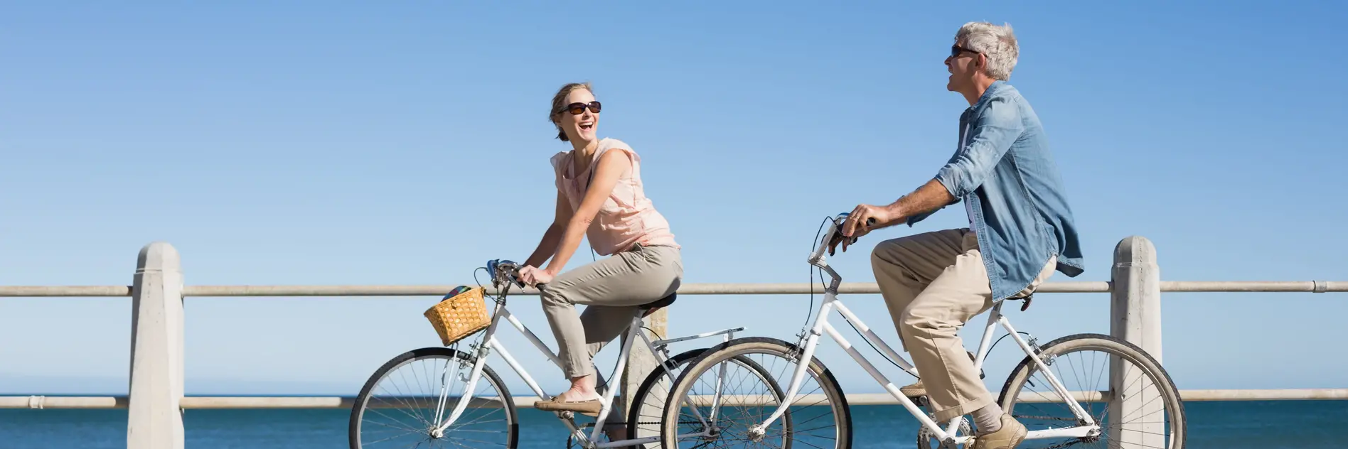 Ein Mann und eine Frau jeweils auf einem weißen Fahrrad fahrend und sich anlachend vor einem Geländer am Meer
