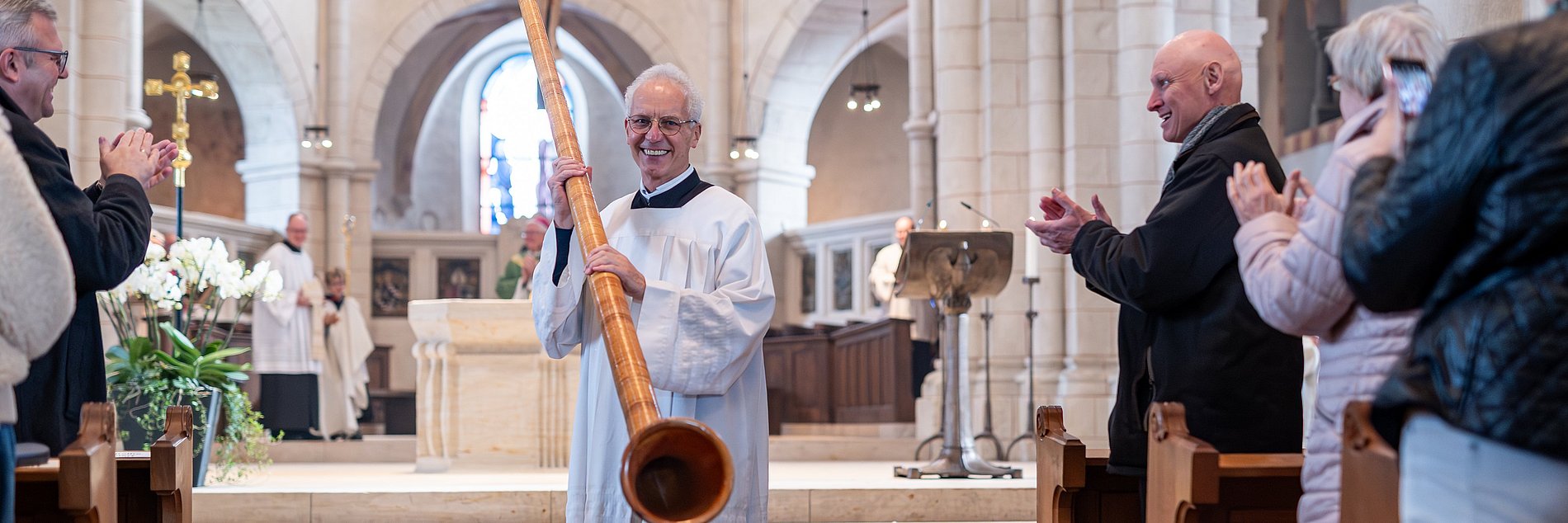 Bruder Elmar Mossbrugger hält sein Alphorn und lächelt, nachdem er im Dom für die Gottesdienstbesucher zum Abschied geblasen hat. Die Gottesdienstbesucher in den Reihen klatschen.