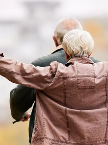 Ein älterer Mann hat seine Frau auf dem Gepäckträger und fährt mit dem Fahrrad - man sieht die beiden von hinten 